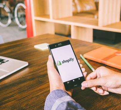 Hands holding a smartphone showing the Shopify logo on a wooden desk, beside a laptop, an orange notebook and a green pen, with shelves and a bicycle behind.