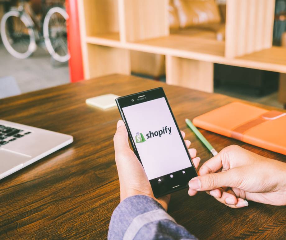 Person holding a smartphone displaying the Shopify logo, seated at a wooden table with a laptop, orange notebook, and a green pen nearby.