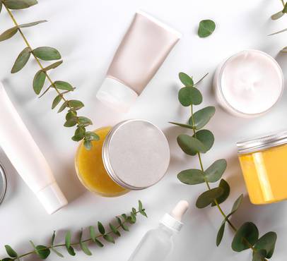 Flat-lay of natural skincare products on a white surface: cream tubes, a dropper bottle, and open jars of golden balm and cream, surrounded by eucalyptus sprigs.