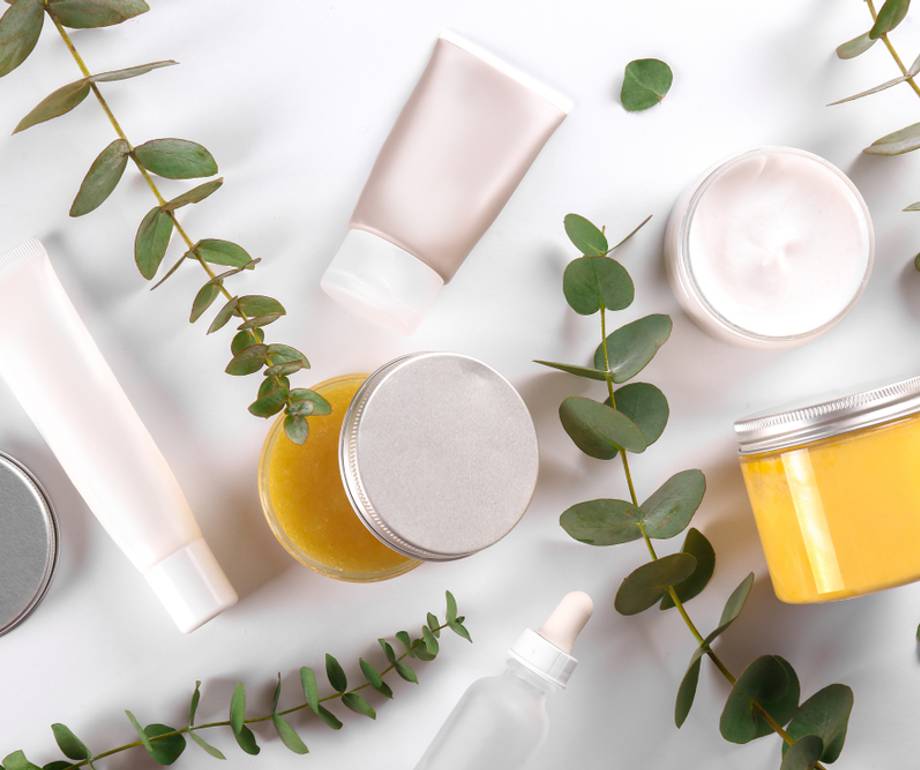 Open cosmetics flat lay with jars, tubes, a dropper, and green eucalyptus leaves on a white background.