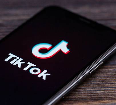 A smartphone with a black screen displaying the TikTok logo in white with cyan and red offset edges, resting on a dark wooden surface.