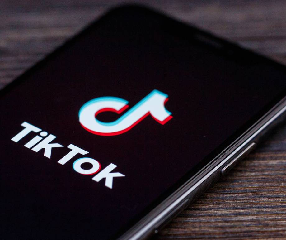A smartphone lying on a wooden surface displays the TikTok app logo on its screen, which is centred in the composition.