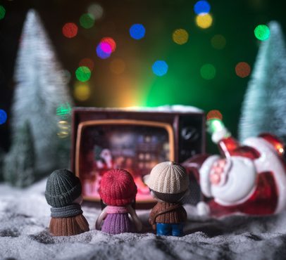 Three small dolls wearing knit hats watch a vintage TV showing a Christmas scene, with a Santa figure lying on its side in front, surrounded by Christmas trees and colourful lights.