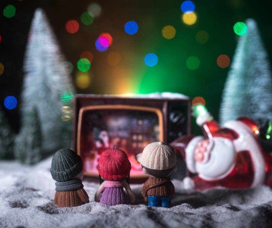 Three small dolls wearing knit hats watch a vintage TV showing a Christmas scene, with a Santa figure lying on its side in front, surrounded by Christmas trees and colourful lights.