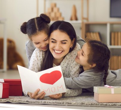 Mother and two daughters lying on the carpet, sharing a joyful moment with a handmade Mother's Day card and gifts, in a cozy living room.