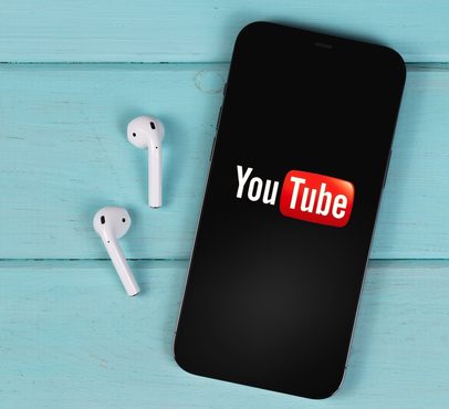 A smartphone displaying the YouTube logo lies on a light blue wooden surface next to a pair of white wireless earbuds.