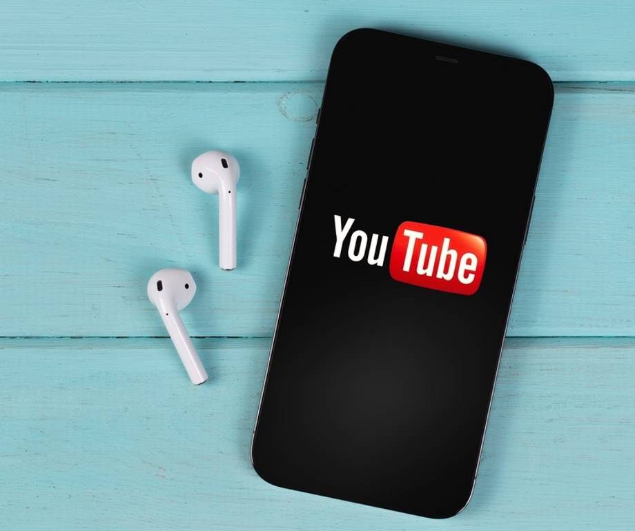 A smartphone displaying the YouTube logo lies on a light blue wooden surface next to a pair of white wireless earbuds.