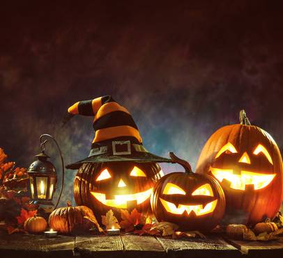 Three illuminated Jack-o'-lanterns with glowing faces, one wearing a witch's hat, surrounded by autumn leaves, pumpkins, and candles on a wooden surface.
