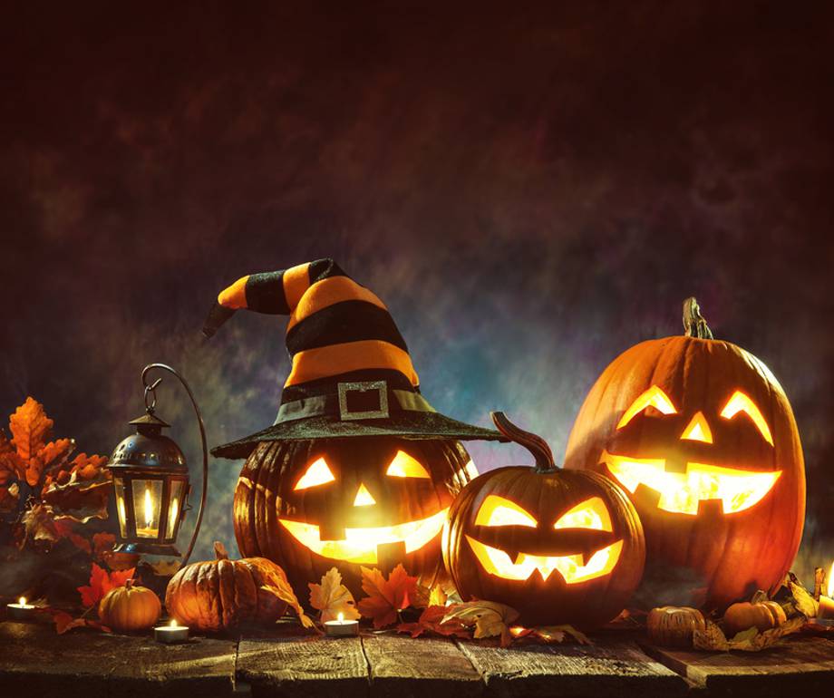 Three illuminated Jack-o'-lanterns with glowing faces, one wearing a witch's hat, surrounded by autumn leaves, pumpkins, and candles on a wooden surface.