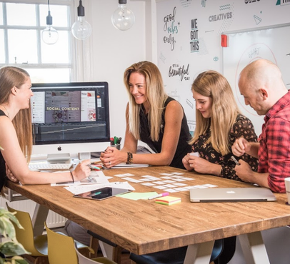 A group of four young adults playing a card game at a wooden table in a modern office space, with a whiteboard and a computer in the background.