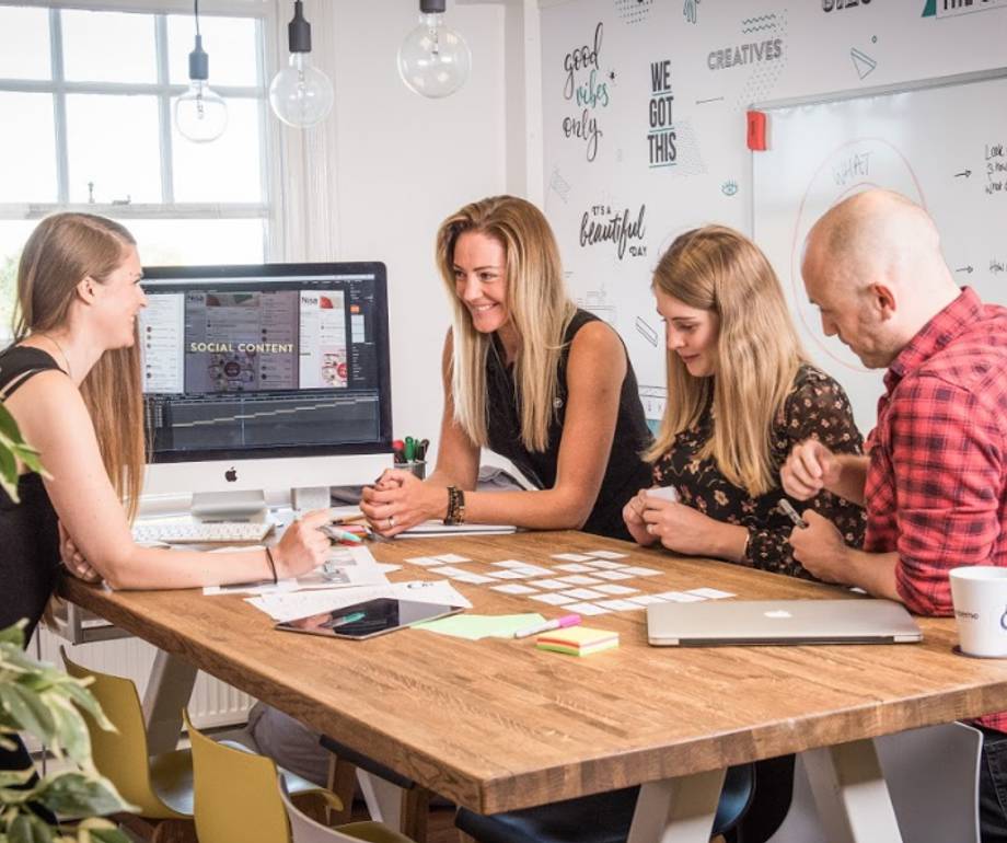 A group of four young adults playing a card game at a wooden table in a modern office space, with a whiteboard and a computer in the background.