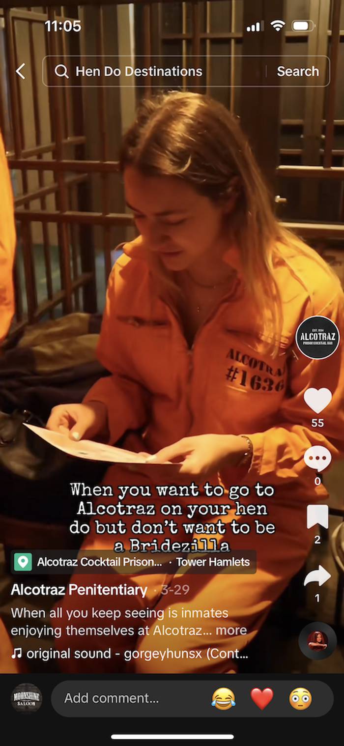 Phone screenshot of a video showing a person with long blonde hair in an orange Alcatraz jumpsuit reading a paper behind bars, with a hen do caption overlaid