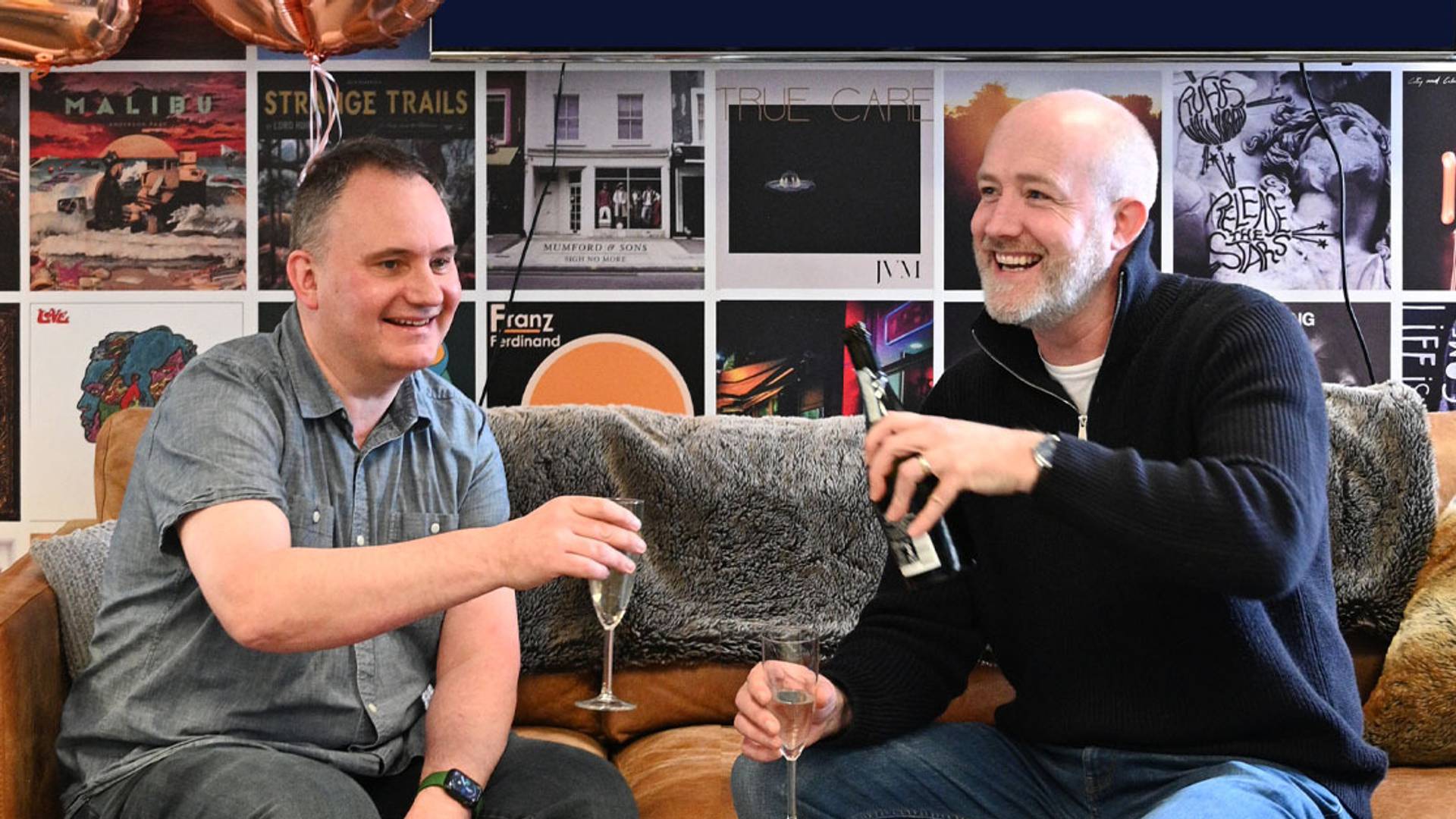 Two men sitting on a sofa, smiling and raising glasses in a toast at an indoor gathering with a wall covered in framed art and posters behind them.