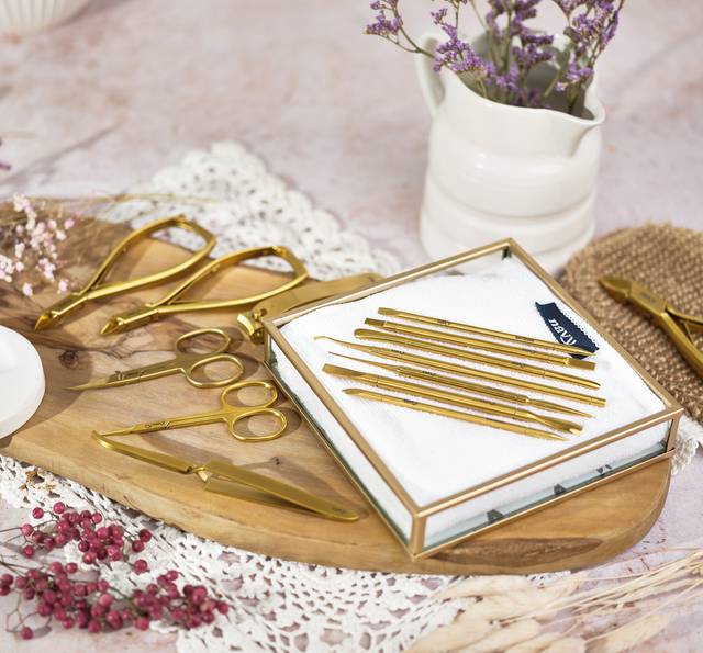 A set of golden manicure tools arranged on a white cloth in a glass box, with additional tools and flowers on a wooden tray, decorated with lace and dried plants.