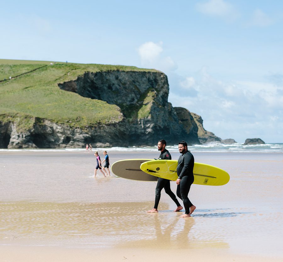Two men in wetsuits with surfboards walk along the sandy beach towards the ocean, with cliffs and a blue sky in the background.