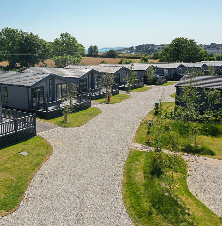 A row of modern, grey cabins with small gardens and gravel paths, surrounded by green trees and open landscape under a clear blue sky.
