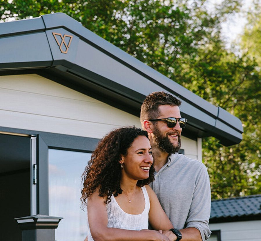 A happy couple leans on a balcony railing outdoors, with a modern mobile home behind them and green trees in the background, enjoying a sunny day.