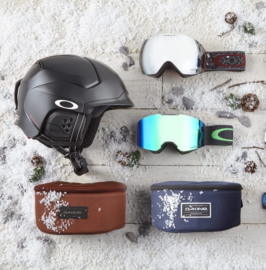 Snowy scene with a ski helmet, two pairs of goggles, and two ski accessories on the snow with pinecones and small branches.