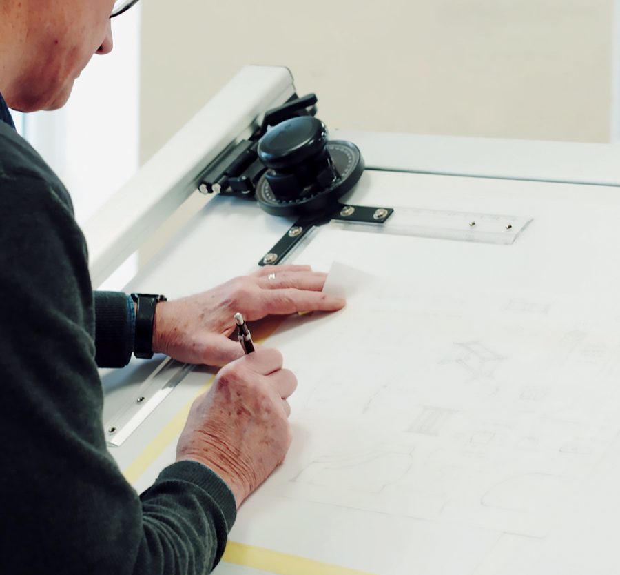 Person drawing on paper with a pen, using a measuring device, at a workspace with a large white surface and a magnifying glass.