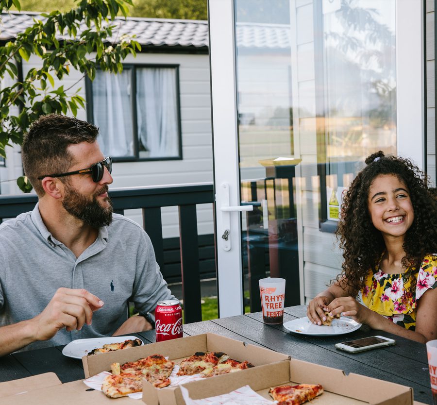 A man with dark hair and beard wearing sunglasses and a grey shirt sits with a young girl with curly hair and a yellow dress, smiling at a backyard pizza gathering.