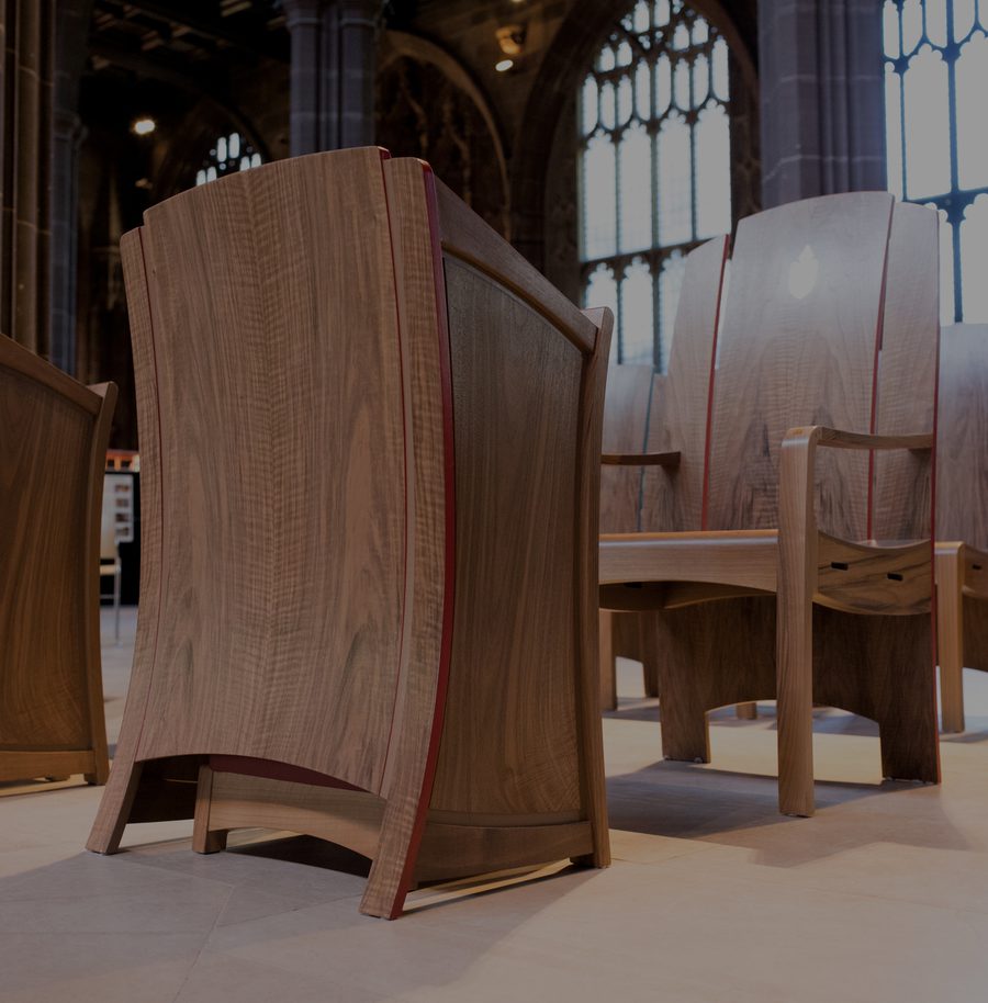 Wooden chairs with curved backs and red edges inside a historic building with tall, arched windows and stained glass.