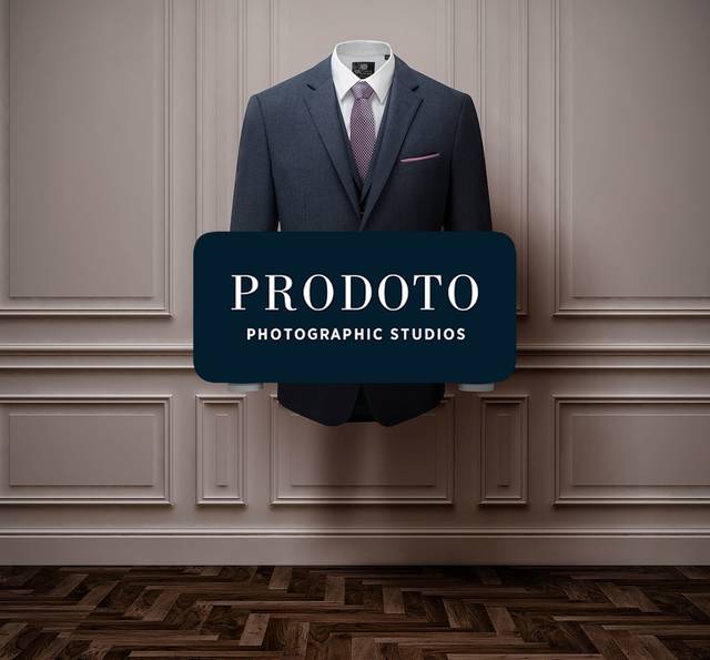 A suit jacket, shirt and tie hang on a wall with decorative paneling, with a "PRODOTO PHOTOGRAPHIC STUDIOS" sign in front.