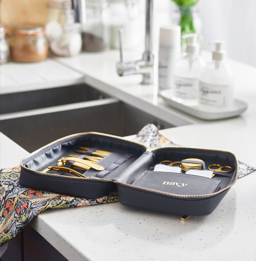 A black toiletry bag with gold scissors, nail clippers, and grooming tools on a colourful cloth, on a kitchen countertop near a sink and soap bottles.