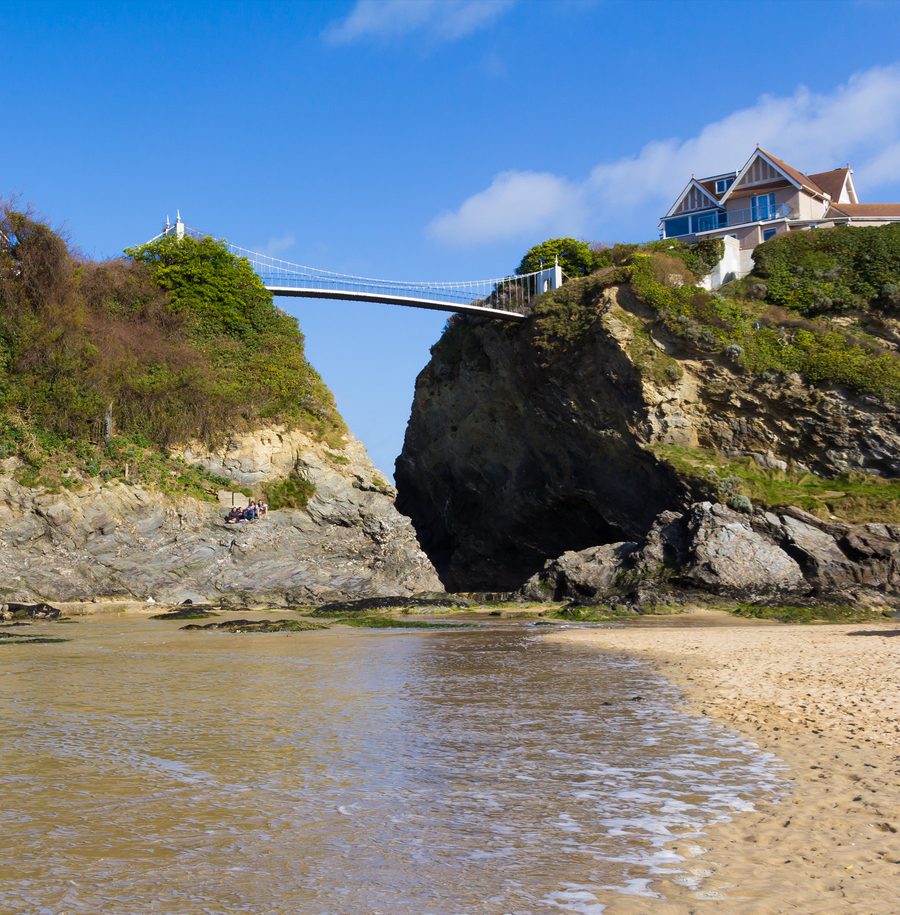 A view of a rocky beach with a bridge connecting two cliffs, and a house on the right top of a hill under a blue sky.