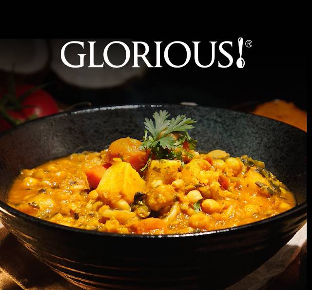 Black bowl of golden vegetable curry with chickpeas, cauliflower and chunks of sweet potato, topped with fresh coriander. White GLORIOUS! logo above.