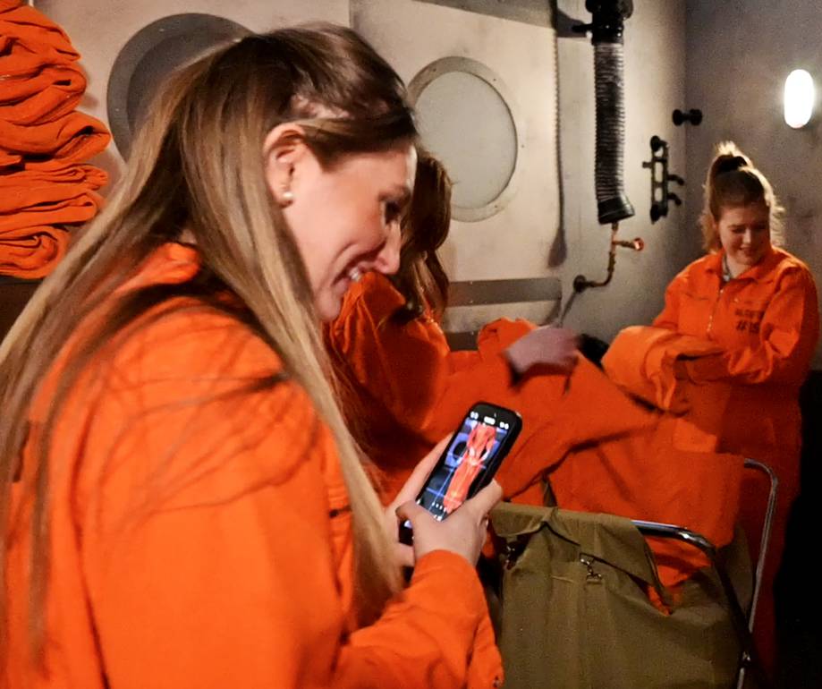 Two smiling women in orange uniforms sit indoors; one looks at her phone, the other turns away, with a wall, pipes, and a light in the background.