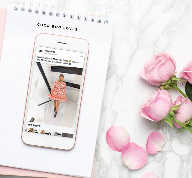 Smartphone displaying a fashion advertisement with a woman in a pink patterned dress, placed on a white notebook and next to pink roses on a marble surface.