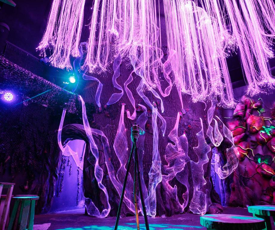 Vivid purple and pink neon lights illuminate an artistic indoor space with flowing fabric sculptures hanging from the ceiling and a textured purple wall in the background.