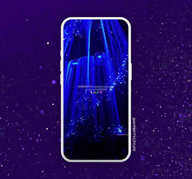 A smartphone displays a blue illuminated water fountain with text about an immersive cocktail experience in London, set against a starry purple background.
