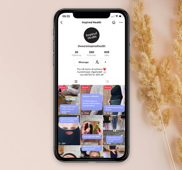 A smartphone displaying an "Inspired Health" social media profile with videos and posts about gut health, set against a neutral background with dried plants.