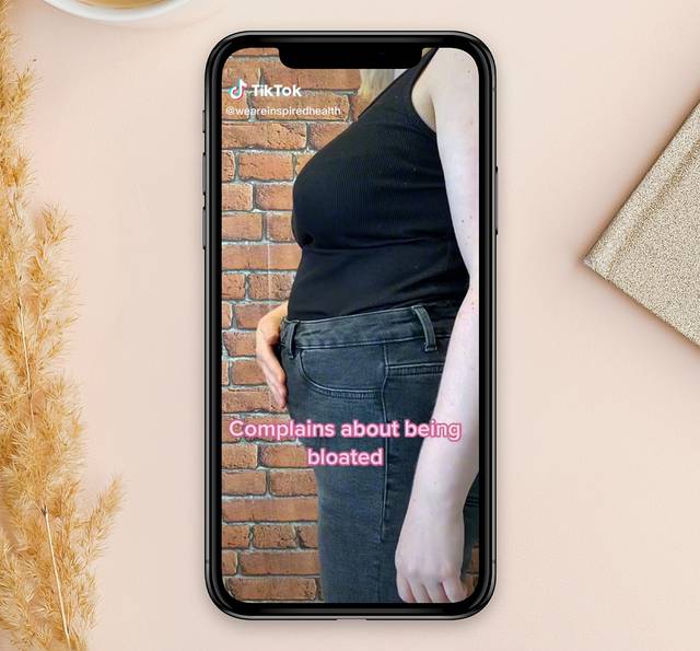A smartphone displaying a side profile of a person with a bloated abdomen, against a brick wall background, with pink text reading "Complains about being bloated".