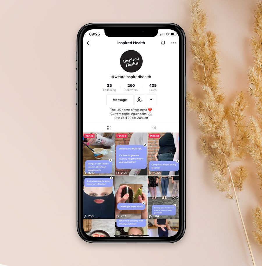 A smartphone displays a social media profile called "Inspired Health" with various videos related to wellness, against a beige background with dried wheat stalks.