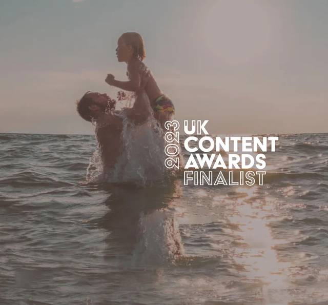 A person lifts a child in swimwear above the sea at sunset, water splashing around them, with white text reading 2023 UK Content Awards Finalist.