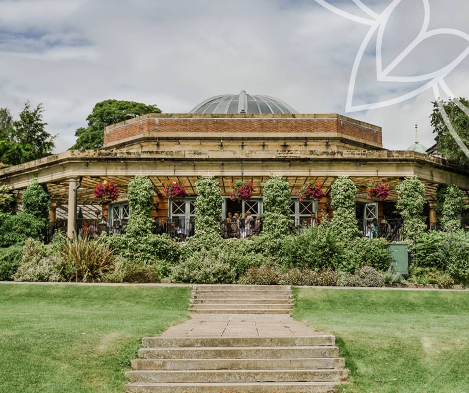 A round building with a glass dome roof, surrounded by greenery and hanging flower baskets, with steps leading up to a terrace.