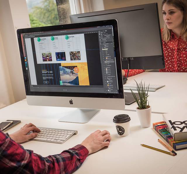 Two women working on a website design project at a white desk with computer monitors, plants, coffee, and stationery in a bright room.