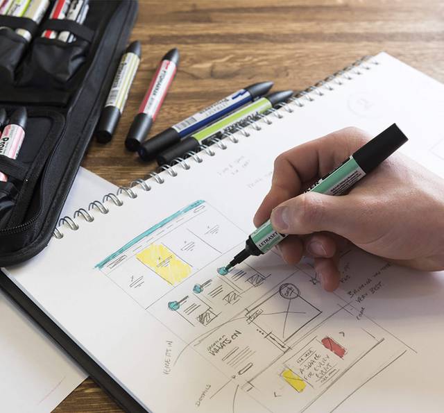 Hand sketching a website wireframe on a spiral notepad with colour-coded annotations, surrounded by markers and a pencil case on a wooden table.