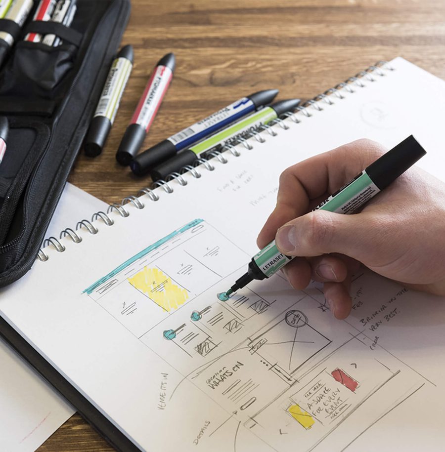 Hand sketching a website wireframe on a spiral notepad with colour-coded annotations, surrounded by markers and a pencil case on a wooden table.
