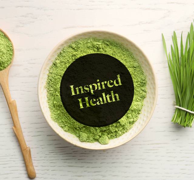 A top-down view of a bowl of green matcha powder with a "Inspired Health" overlay, accompanied by a spoon and a bundle of fresh grass on a white wooden surface.