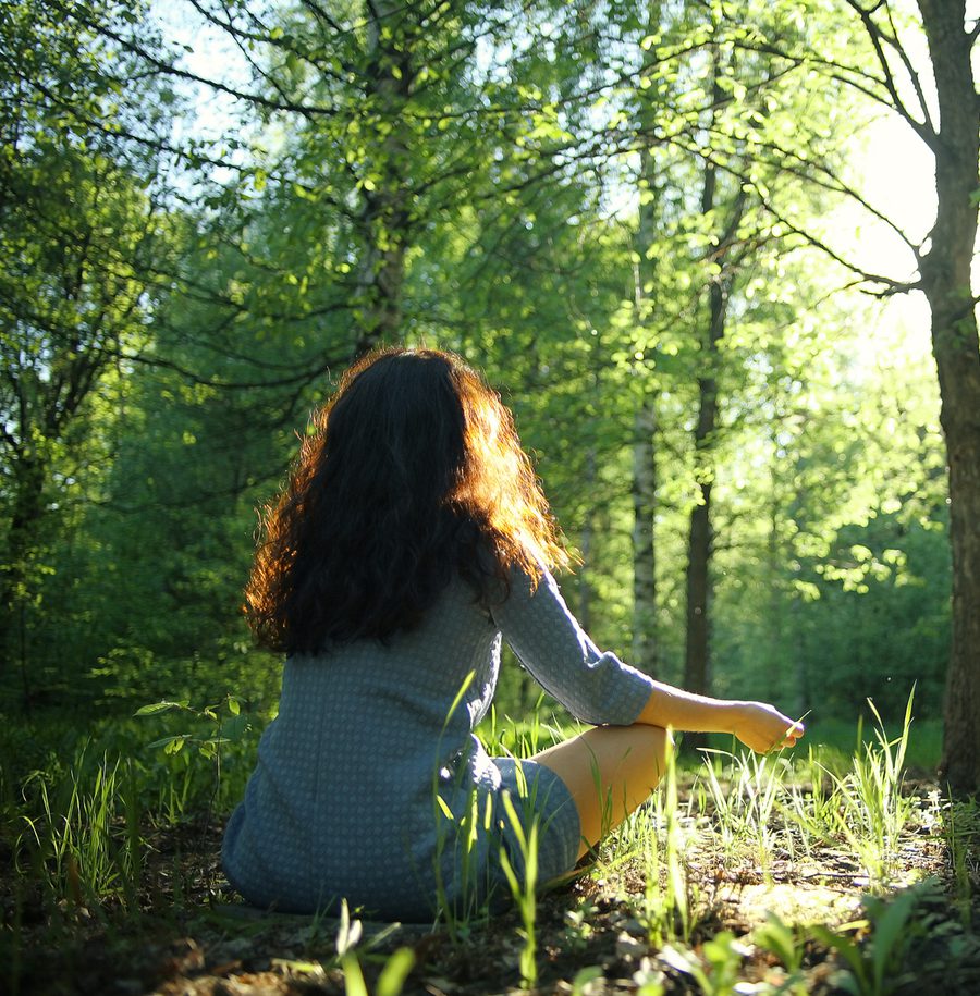 A girl with dark curly hair sitting cross-legged on the forest floor, surrounded by green foliage, during sunlight filtering through the trees.