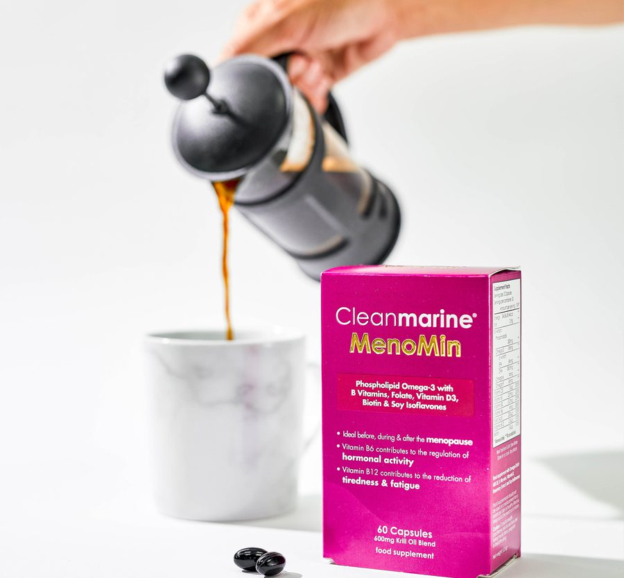 A person pouring dark liquid supplement from a black bottle into a white cup, with a pink box of Cleanmarine MenoMin supplements in the foreground.