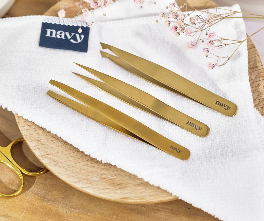Three gold tweezers on a white cloth with a navy logo, placed on a wooden surface, with pink dried flowers nearby.