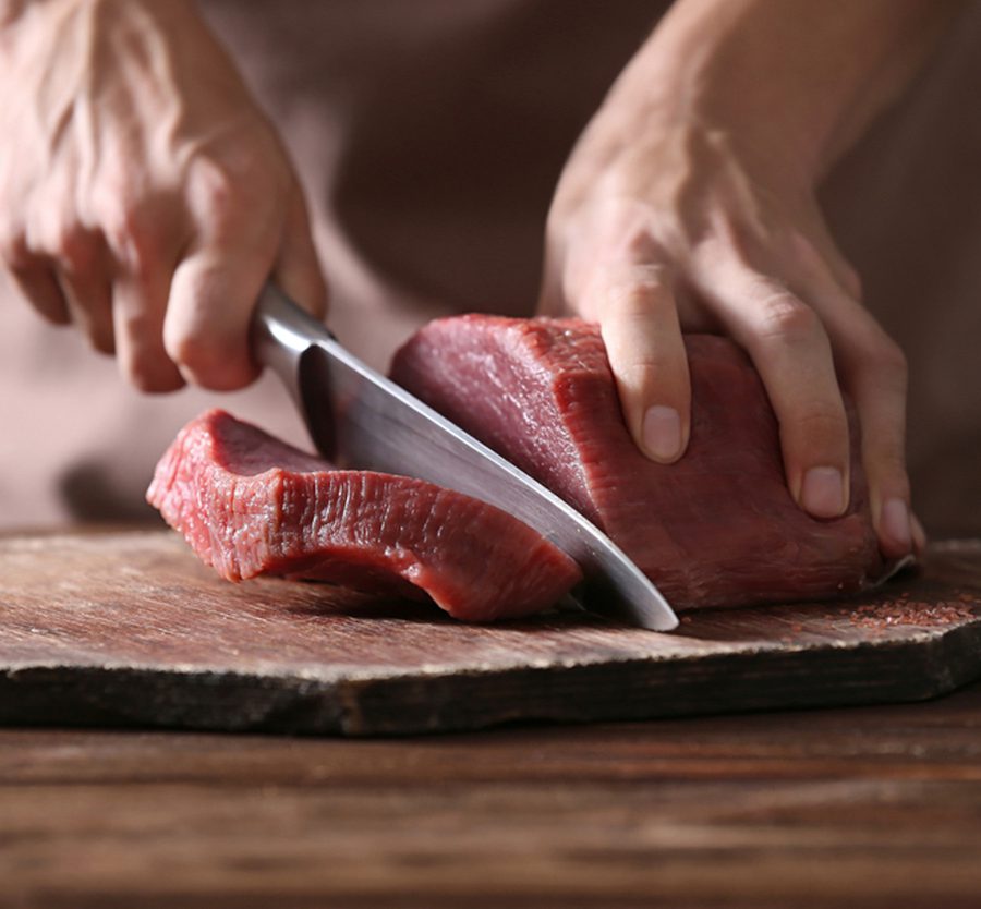 Hands slicing raw meat on a wooden cutting board, focusing on the knife and meat with a blurred background.