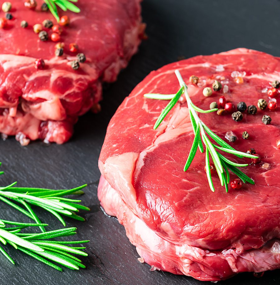 Raw beef steaks garnished with black peppercorns and rosemary sprigs on a dark surface.