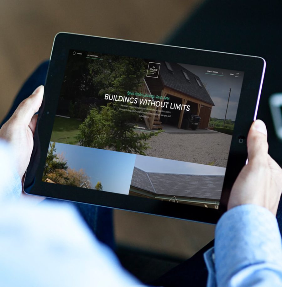 Person holding a tablet displaying a website about buildings, with a focus on a house and surrounding greenery in the background.