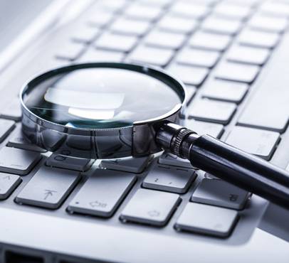 A magnifying glass with a black handle rests on a silver laptop keyboard, its lens showing reflections, with keys in soft blue-grey light.