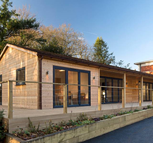 A modern wooden house with large glass doors and windows, a small balcony with a wooden railing, set against a background of trees and a clear blue sky.